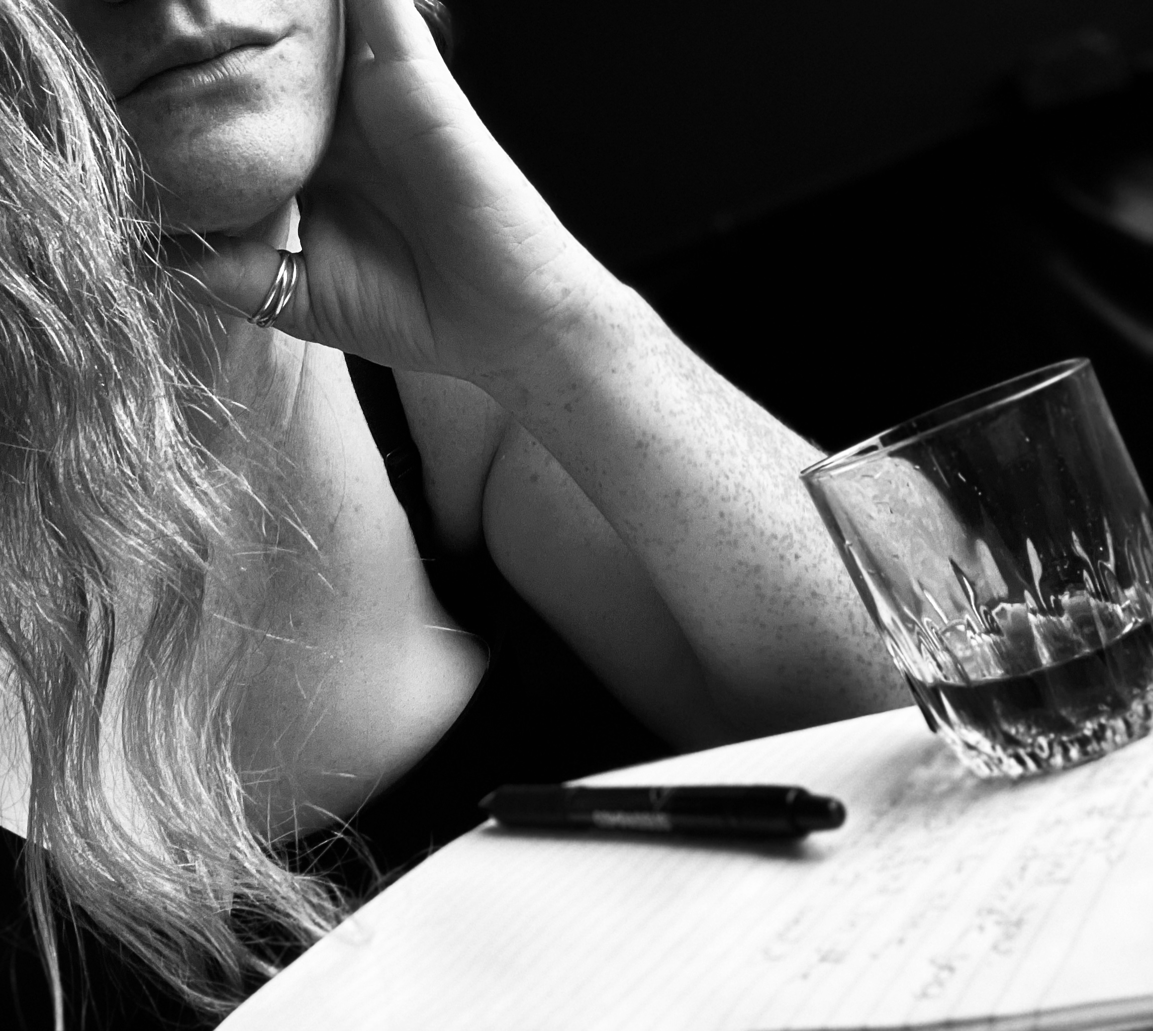 Picture of a woman laying on a bed with a notpad and pen with her long hair swept in front of her on one side and her hand holding her face on the other. you can only see up to her lips and doesn't show her whole face. This woman is writing, contemplating and sipping on vanilla bean vodka. this image holds the link that leads to the "Erotica" page