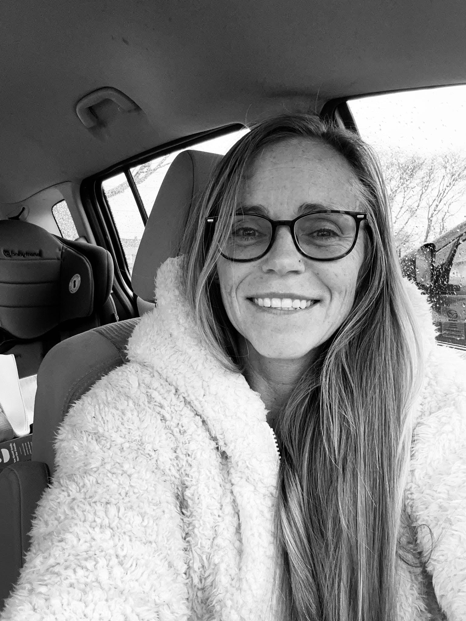 BLACK AND WHITE PICTURE OF A WOMAN SITTING IN HER CAR TAKING A SELFIE SMILING AND OPEN TO HELPING OTHERS WITH LIFE COACHING IN A WHITE FLUFFY COAT